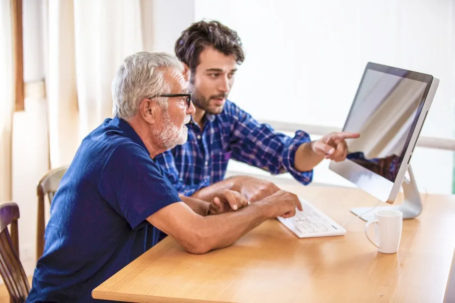 In-Home Computer Repair in progress with older man learning to use his computer at his home office desk while a computer repair technician in a blue buttondown shirt and lanyard is showing him how to accomplish the task.